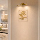 Gold Hebrew letter wall art mounted on a beige textured wall, illuminated by a gold wall sconce. The acrylic plaque is shown in a modern, elegant living room setting.