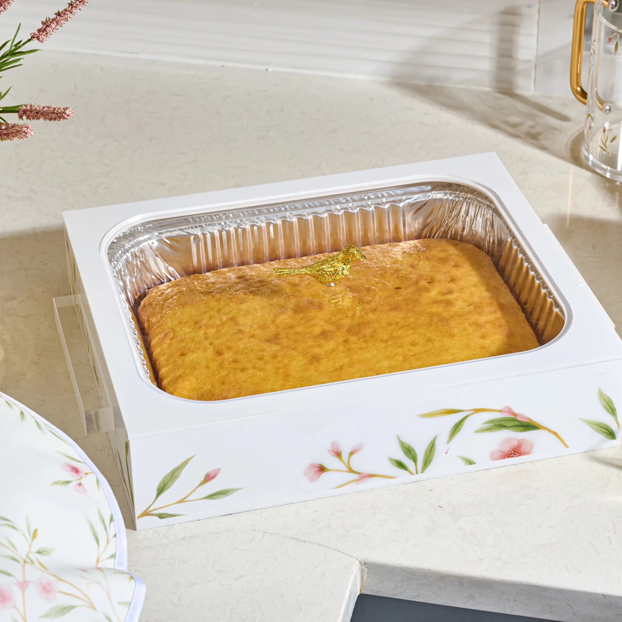 A rectangular golden-brown baked cake or casserole presented in a white floral acrylic tray with a silver foil-lined bottom, set on a light countertop.