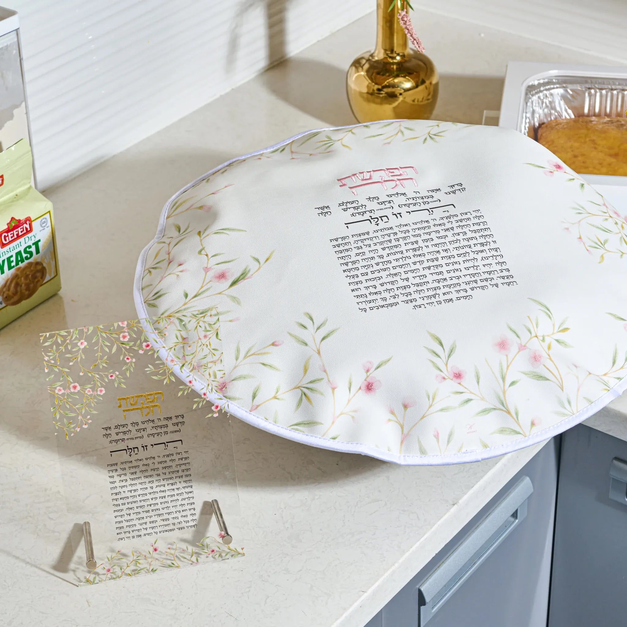 A floarl printed card with floral and leaf patterns in soft green, pink, and gold, featuring Hebrew text at the top and a block of smaller text below, displayed on a clear stand in a kitchen setting.