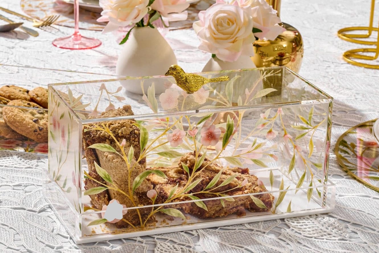 Clear decorative lucite cake dome with greenery and a gold bird on a table with cookies and flowers.