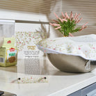 A decorative Challah dough cover over with Hebrew text and a floral border stands on a kitchen counter next to a box of Leavened yeast. In the background, a white electric blanket with a matching floral pattern is visible, along with a vase of pink flowers and window blinds.