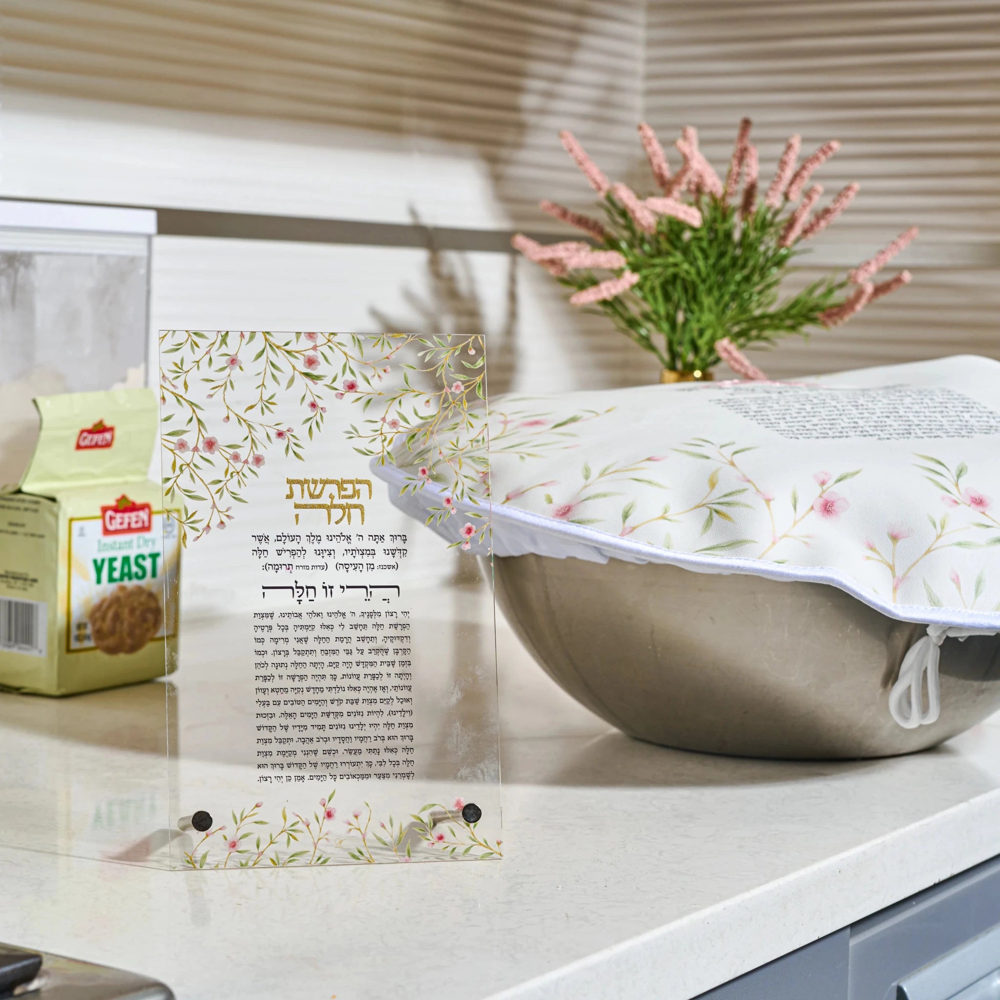 A decorative Challah dough cover over with Hebrew text and a floral border stands on a kitchen counter next to a box of Leavened yeast. In the background, a white electric blanket with a matching floral pattern is visible, along with a vase of pink flowers and window blinds.