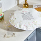A round white floral challah dough cover with a floral border and printed Hebrew text, displayed on a kitchen countertop next to a  clear acrylic cup and a box of yeast.