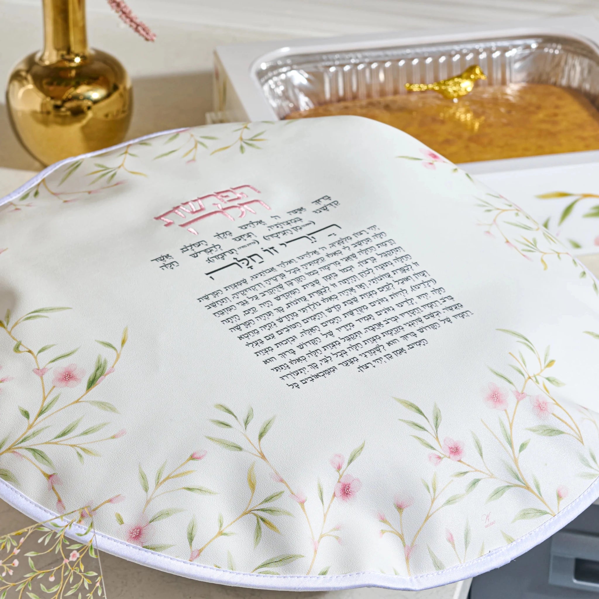 A round white Floral  Chsllsh  cover with printed Hebrew text and pink floral illustrations, shown on a table next to a gold-colored decorative bottle and a foil pan of leavened bread.
