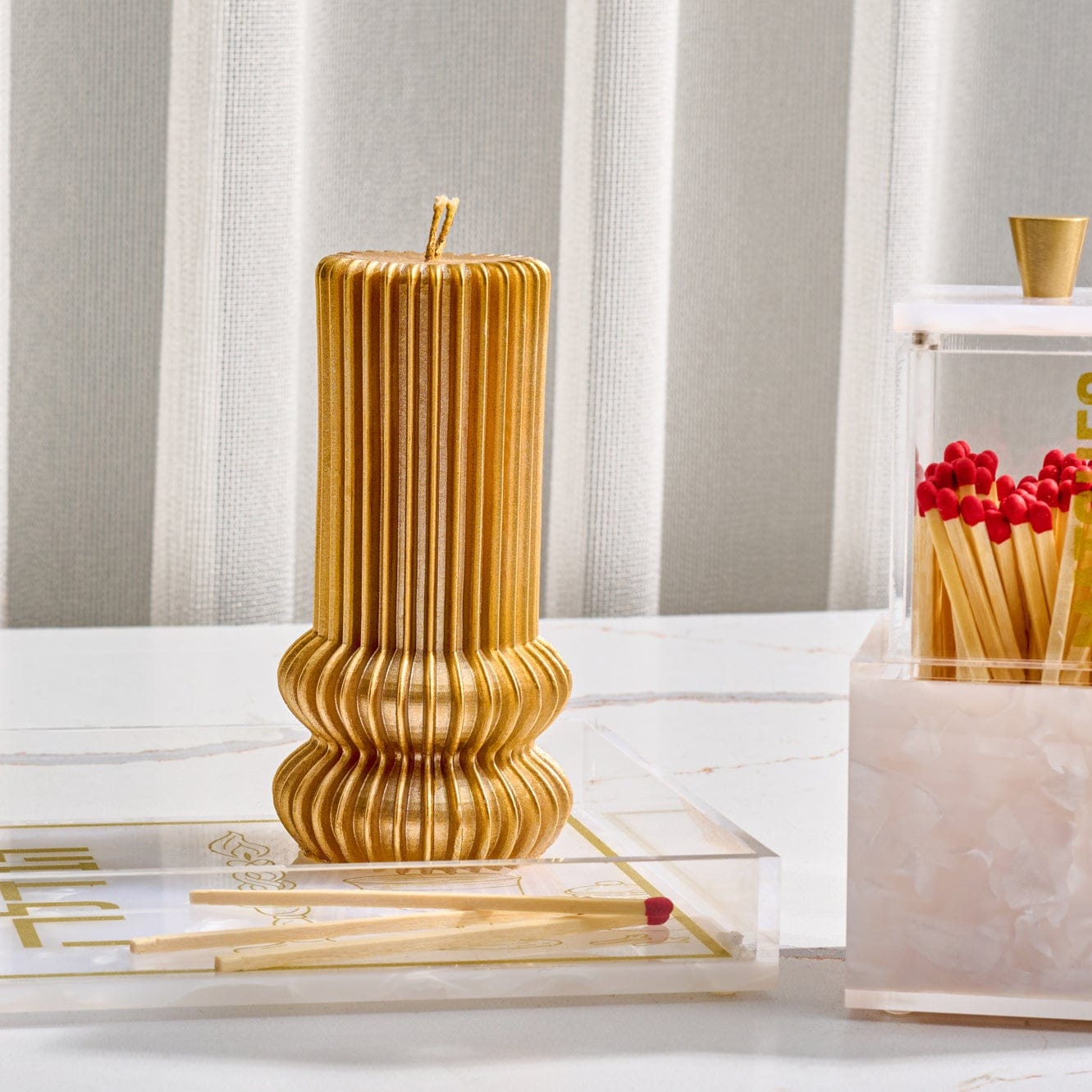 Fluted Havdalah candle in metallic gold finish featuring sculpted ribbed design and double wick, displayed on clear acrylic tray with matches, elegant modern Judaica setting.