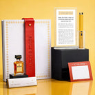 Luxury Mishloach Manot gift box featuring a gold-bordered Classic 2.0 Hadlokas Neiros Block With Lighter in a lucite stand, a white leatherette pouch with a red strap, and a mini Disaronno liqueur.