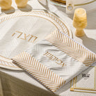 Herringbone Afikoman bag in cream with gold striped accents and Hebrew “Afikoman” lettering, displayed on a matching Seder plate at a Pesach table.