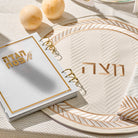 Herringbone matzah cover in cream with gold accents and Hebrew “Matzah” lettering, displayed on a coordinating Seder plate.