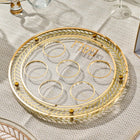 Herringbone Seder plate with clear lucite wells and gold-tone metal rim, displayed on a patterned tablecloth for Passover.