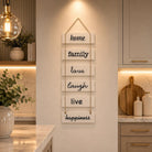 A vertical wooden sign with six white rectangular panels hanging by rope, each reading a positive word: home, family, love, laugh, live, happiness. The sign is displayed on a warm beige wall in a modern kitchen setting.