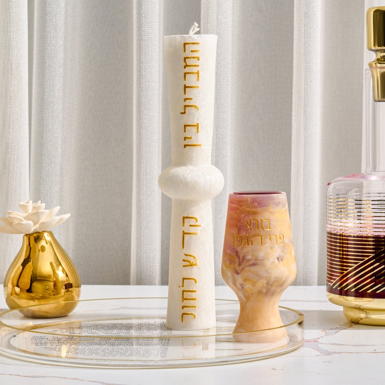 Hourglass Havdalah candle in matte white wax with sculpted center detail and gold Hebrew lettering, displayed on a clear round tray beside a marbled Kiddush cup and modern Judaica décor.