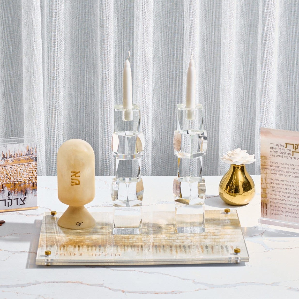 Jerusalem taupe hadlokas neiros set featuring tall crystal candle holders on a modern acrylic tray with a western wall stone pattern alongside a matching matchbox and tzedakah box on a marble table.