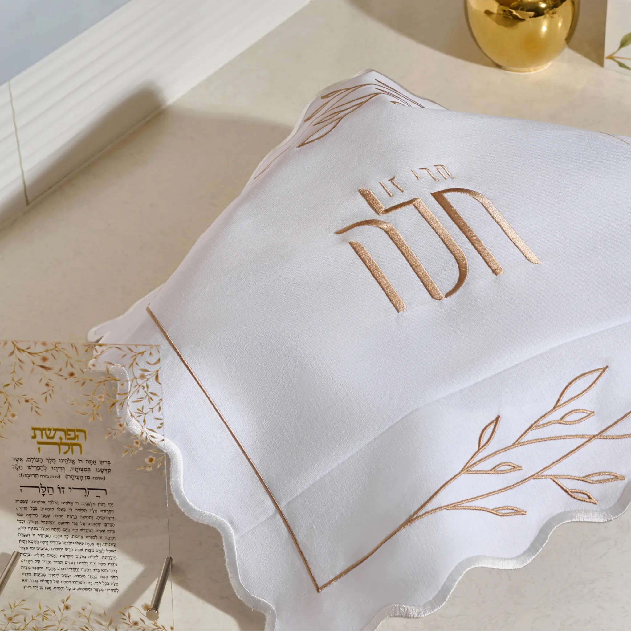 A white  linendecorative challah cover with a scalloped edge and a rose gold embroidered design featuring Hebrew text and leafy accents, set against a plain white background.