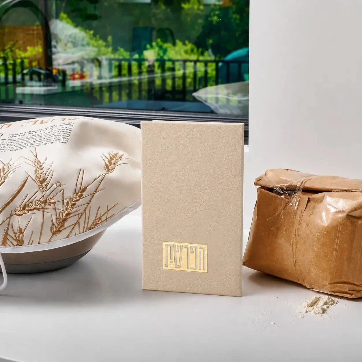 Tan leather Hafrashas Challah booklet displayed with dried wheat and kraft packaging on white surface near window.