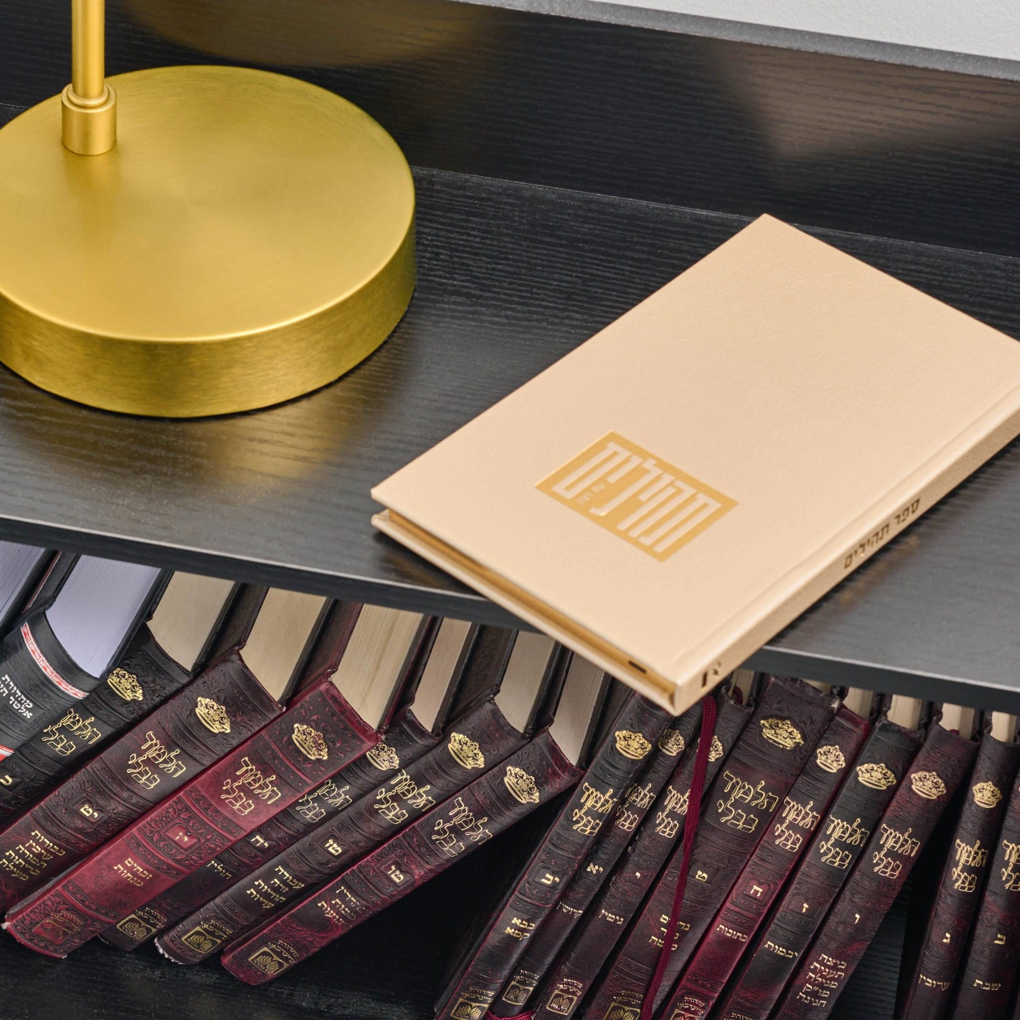 A gold  hardcover tehillim book with a gold logo lies on a dark bookshelf next to a gold lamp base, with rows of dark leather-bound books underneath.