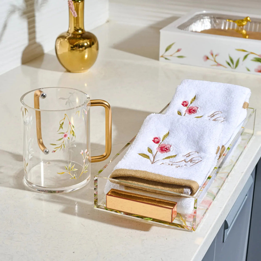 A styled tabletop scene with a clear acrylic tray holding folded white embroidered towels and a gold-toned soap bar, alongside a clear glass mug with a gold handle and floral print. A gold vase and a floral-patterned box are also visible in the background on a light countertop.