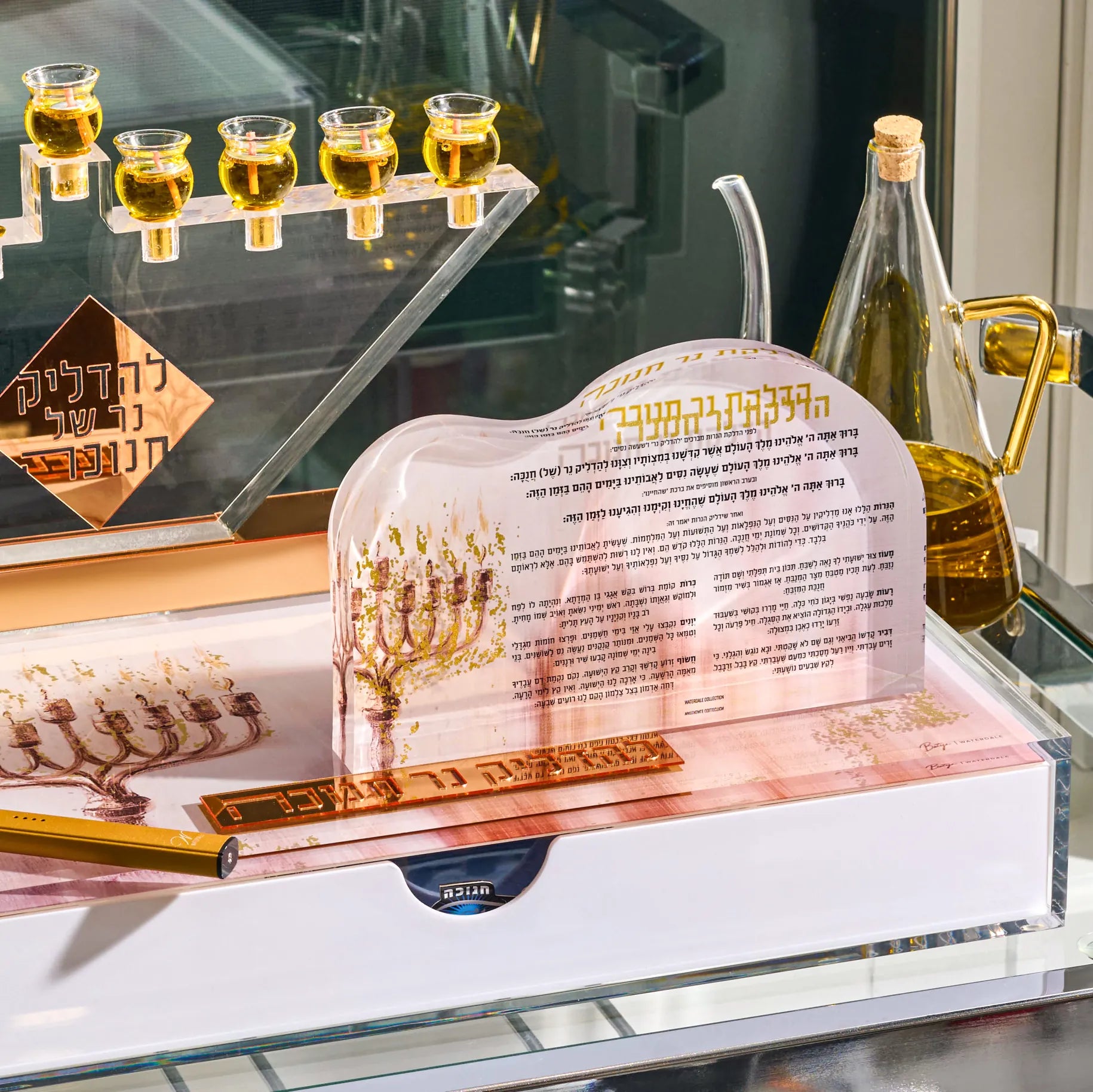 A Hanukkah menorah set displayed on a glass shelf, featuring a clear acrylic menorah with a printed menorah illustration and Hebrew text, alongside a gold-colored metal menorah, a gold candlestick, a glass bottle of olive oil, and small glass cups.