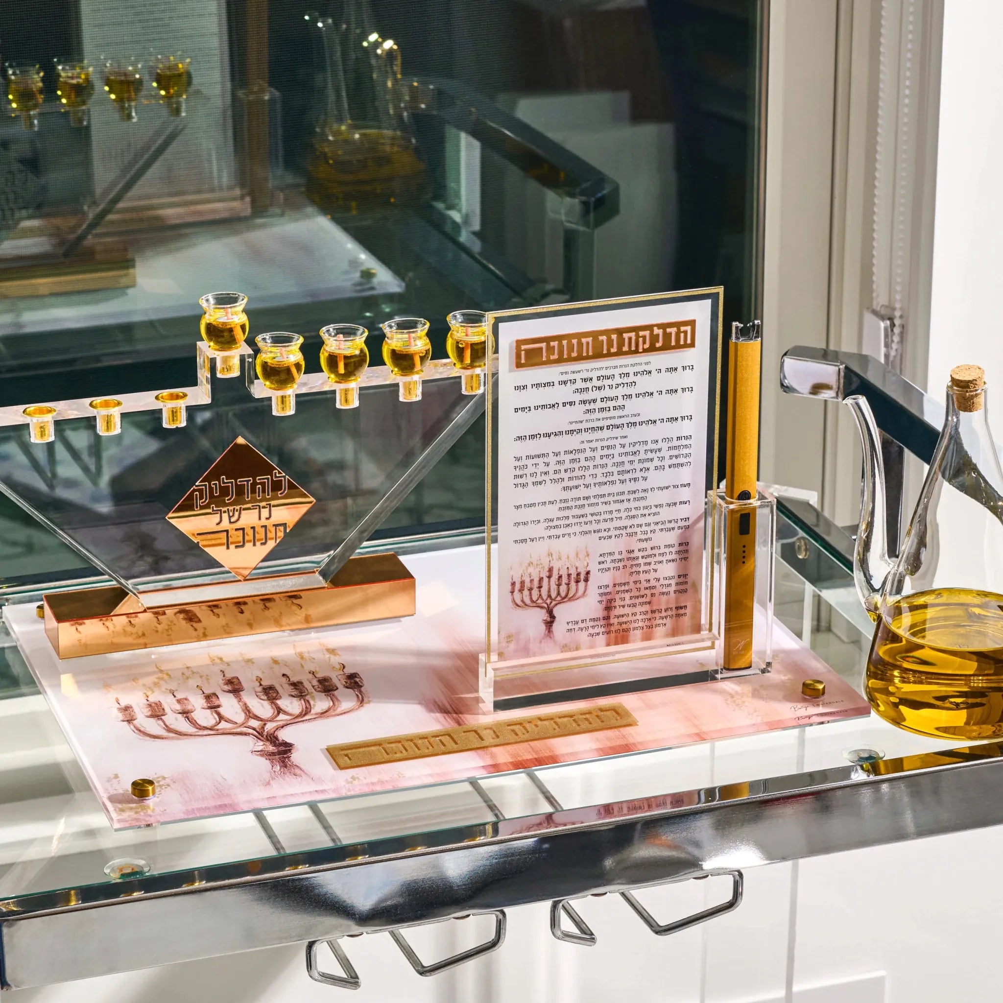 A polished silver and gold Hanukkah menorah display with clear acrylic cups filled with oil, shown on a glass shelf beside a glass pitcher and an informational plaque in Hebrew.