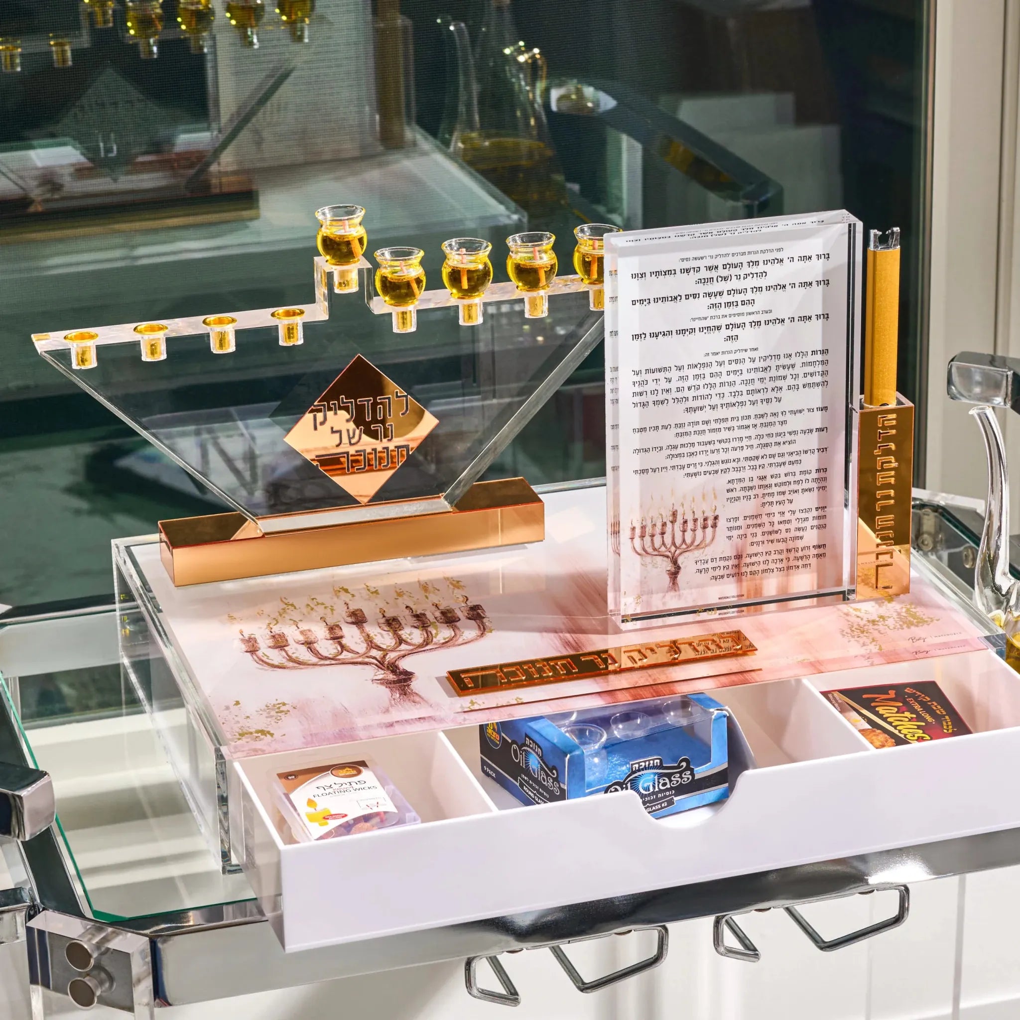 A Hanukkah menorah set displayed on a glass shelf, featuring a clear acrylic menorah with gold-toned cups and base, a printed instruction card, a gold candle, and a white drawer containing candles , glasses...