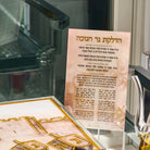 A printed Hebrew brachos card displayed on a clear stand, with a decorative gold-toned plaque or box in the foreground and a blurred indoor setting behind it.