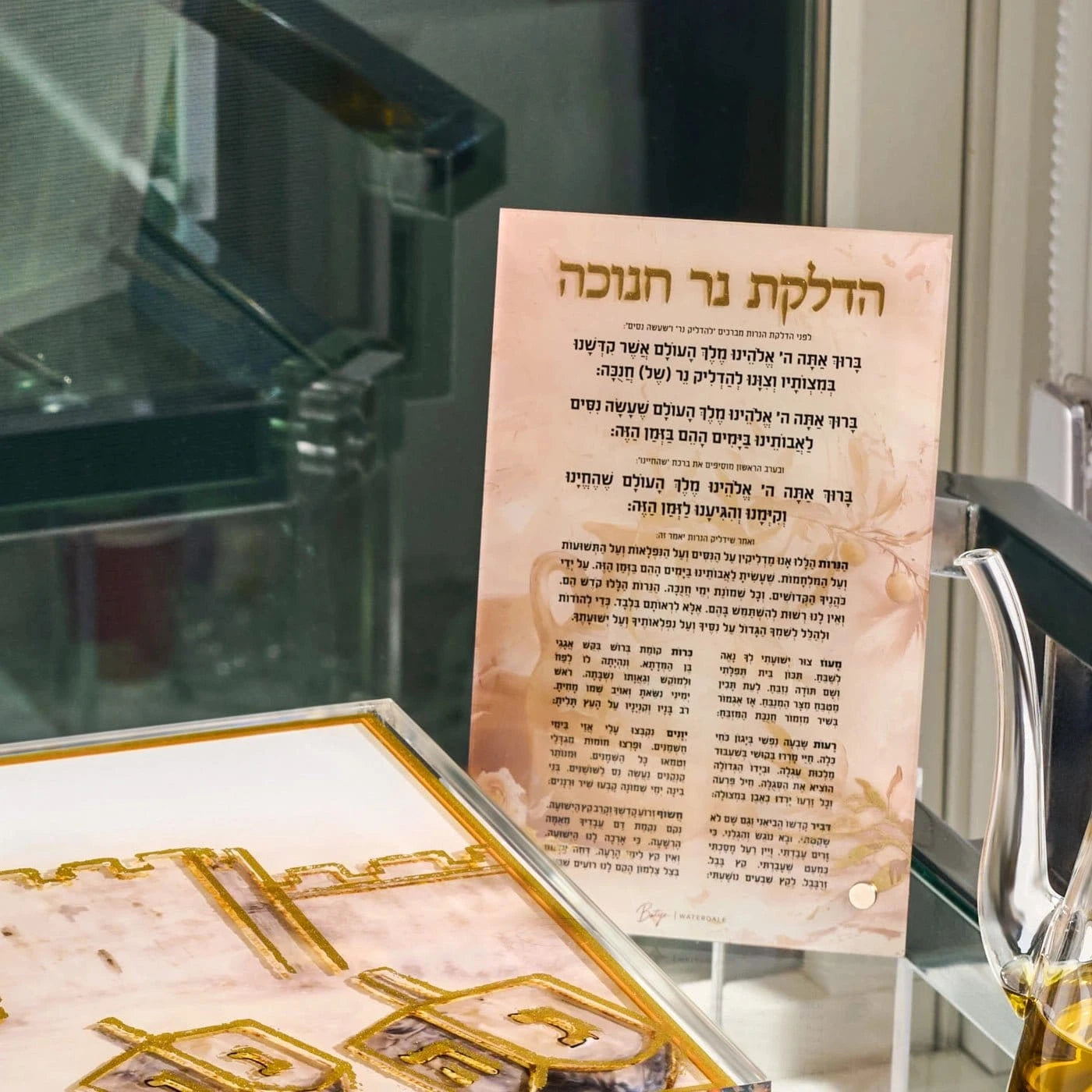 A printed Hebrew brachos card displayed on a clear stand, with a decorative gold-toned plaque or box in the foreground and a blurred indoor setting behind it.