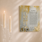 A square wall art print with Hebrew text and an abstract gold, blue, and white design hangs on a cream wall above a marble console table styled with lit candles, a small candle holder, and white flowers.