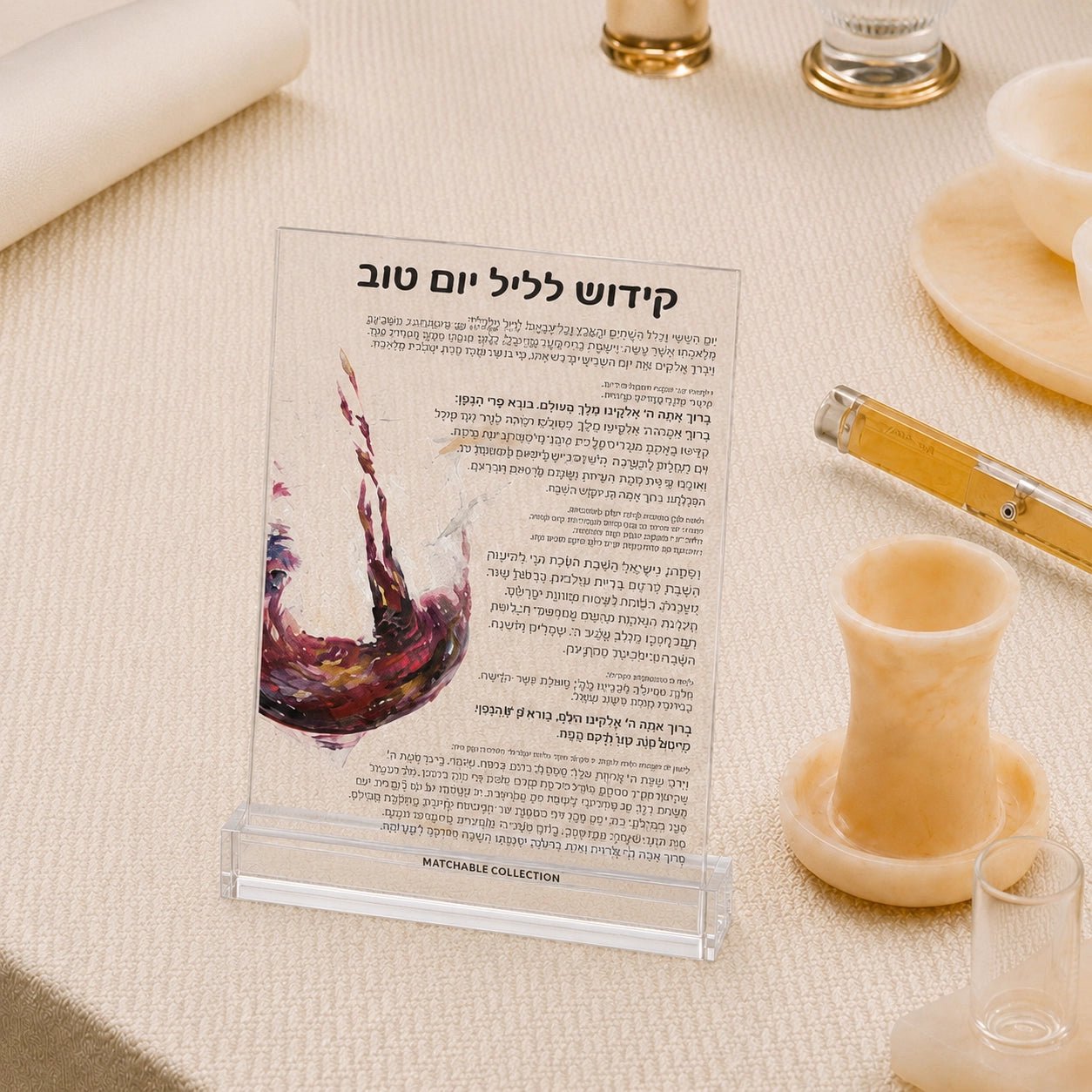 A clear acrylic sign holder displaying a Hebrew wine card with a red wine splash illustration, set on a beige tablecloth with dining props like a glass, candle, and plates.