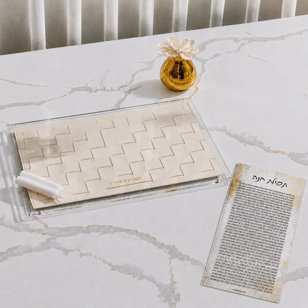 A hadlokas neiros  tray set on a white marble table, featuring a clear acrylic base with a beige woven basket insert, two white candles, a small gold decorative vase, and a printed Hebrew tefilas chana card