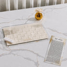 A hadlokas neiros  tray set on a white marble table, featuring a clear acrylic base with a beige woven basket insert, two white candles, a small gold decorative vase, and a printed Hebrew tefilas chana card