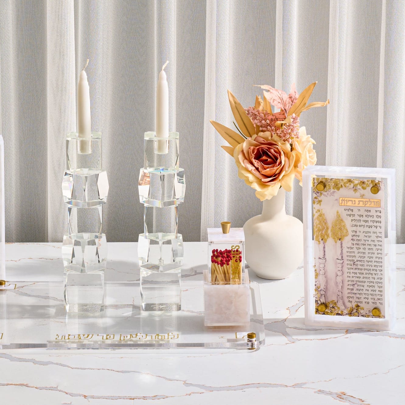 Pearlstone hadlokas neiros set with crystal candle holders an acrylic matchbox and prayer card on a crushed marble and acrylic tray with gold Hebrew text and floral accents on a marble table.