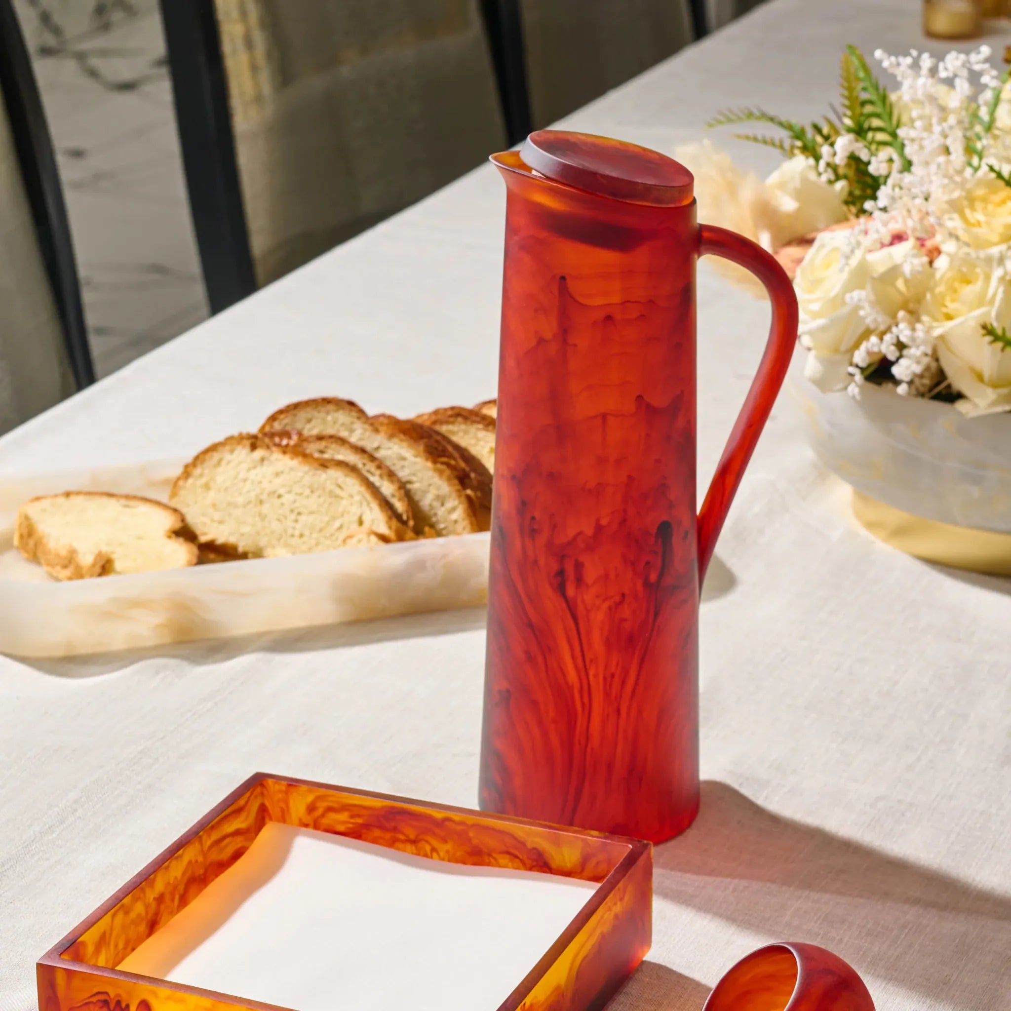 A translucent amber-resin glass pitcher with a matching square bowl and small spoon, styled on a white tablecloth with bread and flowers in the background.
