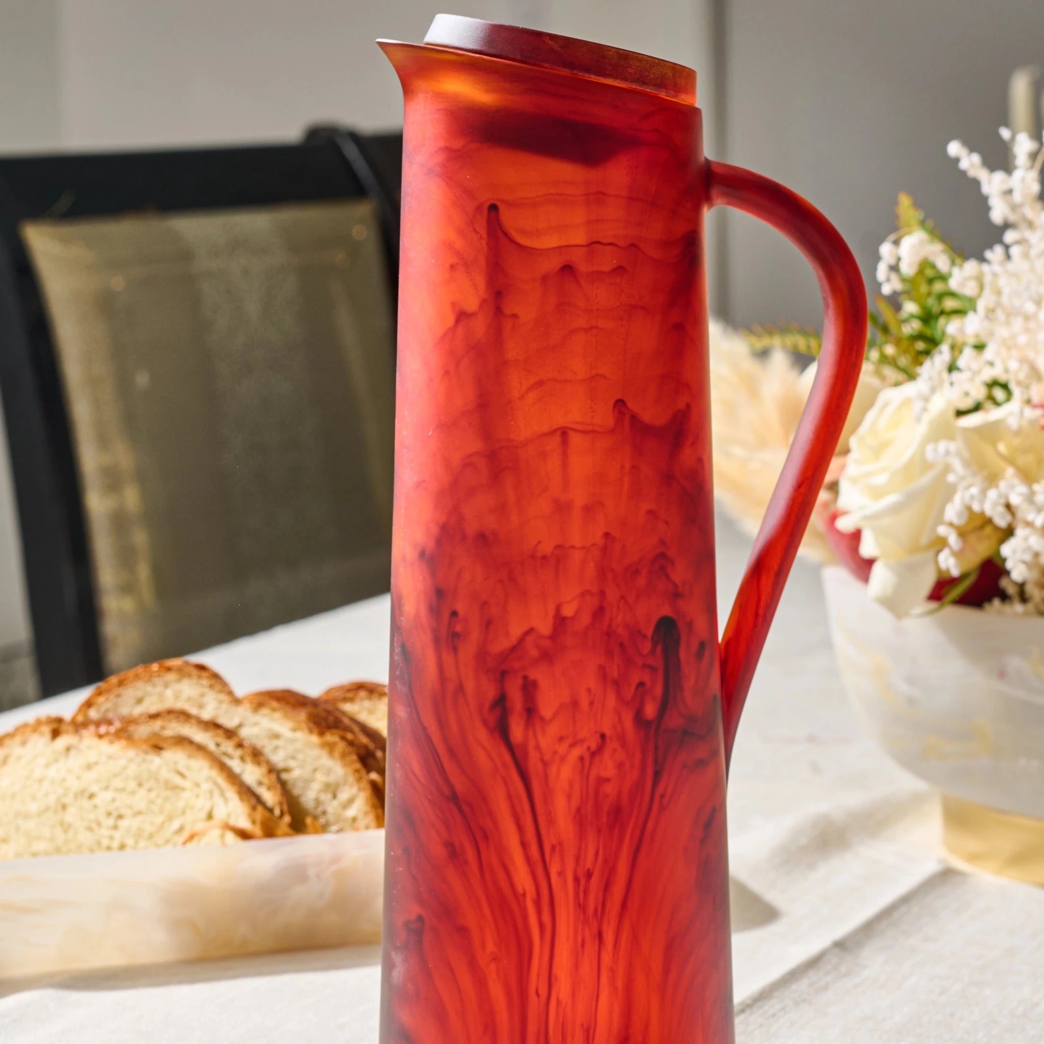 A tall translucent amber resin pitcher with a marbled swirl pattern, set on a dining table with bread and flowers in the background.