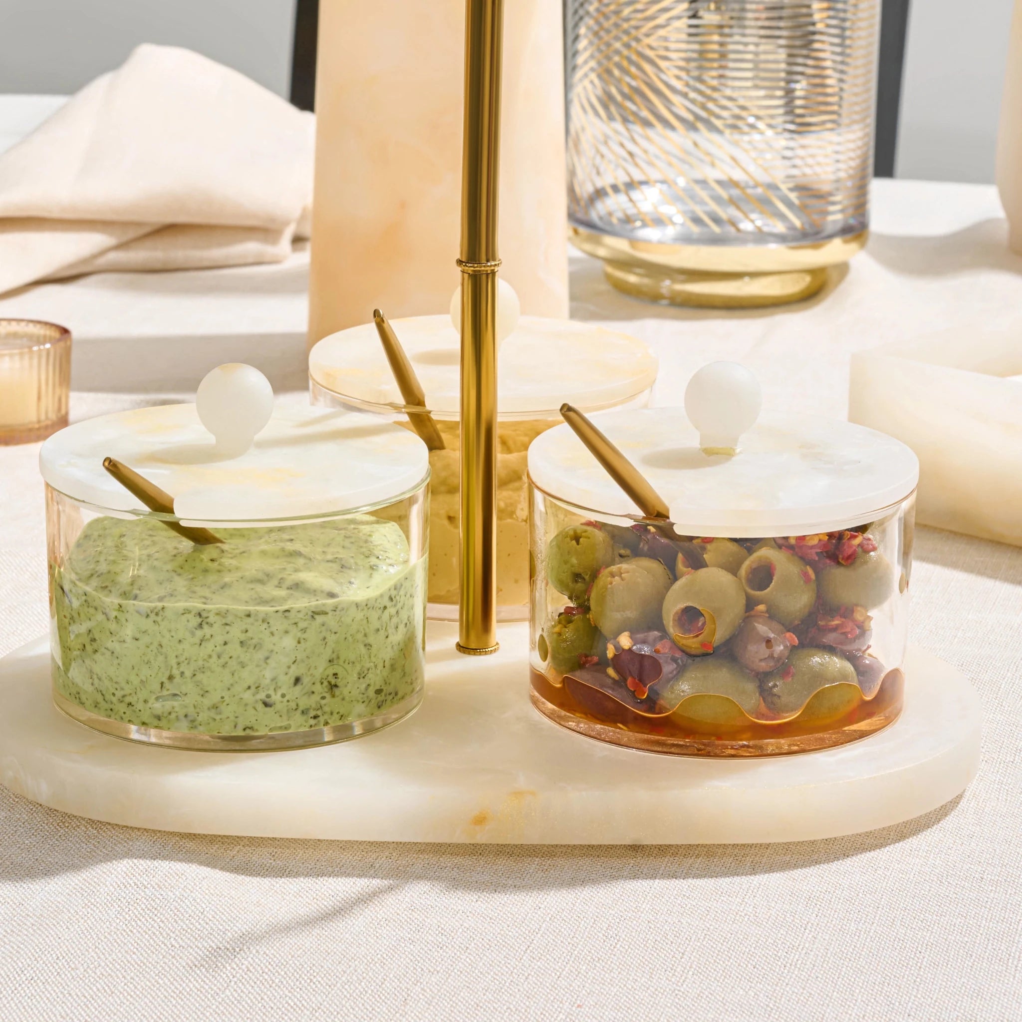 Three clear glass condiment jars with resin lids and gold spoons, arranged on a round roesin  tray. The jars contain green herb spread, a yellow spread, and sliced olives with red peppers, set on a beige tablecloth with dining props in the background.