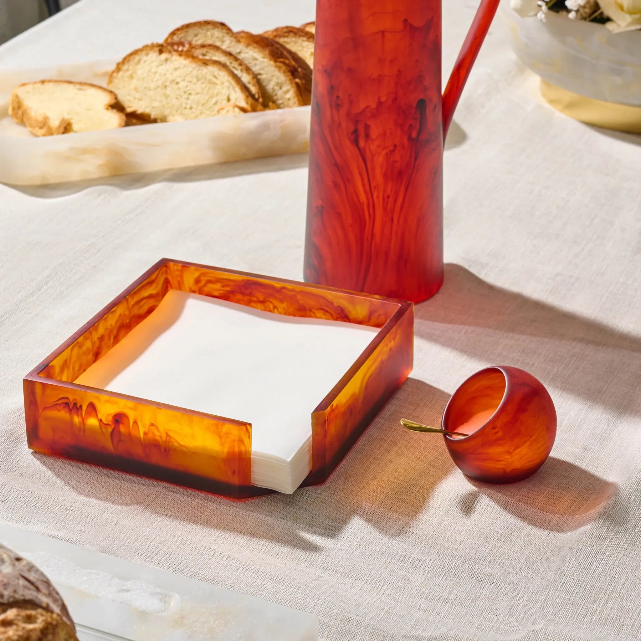A set of amber resin  tableware on a white tablecloth, including a square napkin holder with white napkins, a tall resin pitcher , and a small salt cellar 