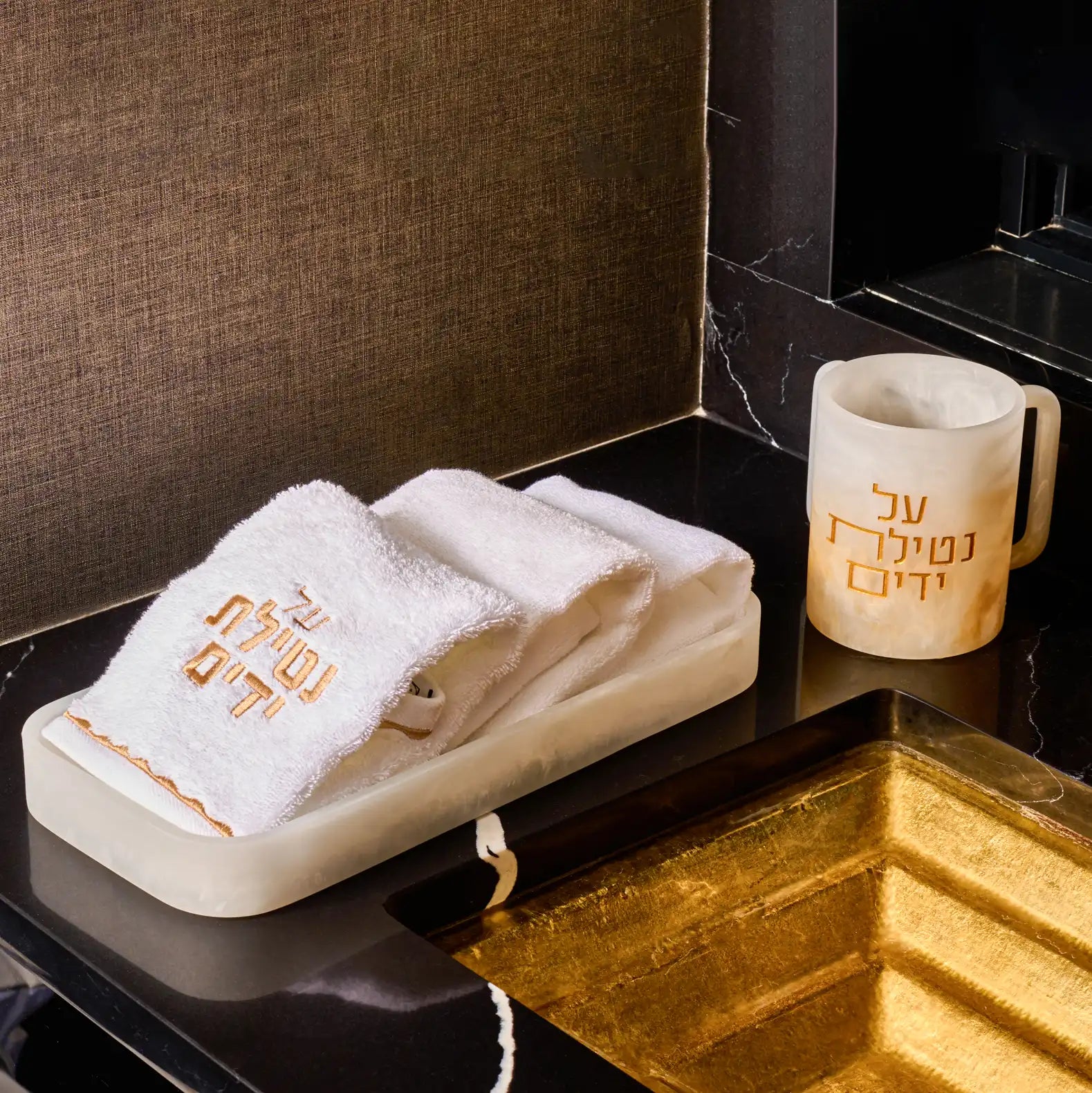 Ombre resin washing cup with gold “Al Netilas Yadayim” Hebrew lettering, displayed beside a white embroidered hand towel on a resin towel tray on a sleek black marble countertop near a modern sink.