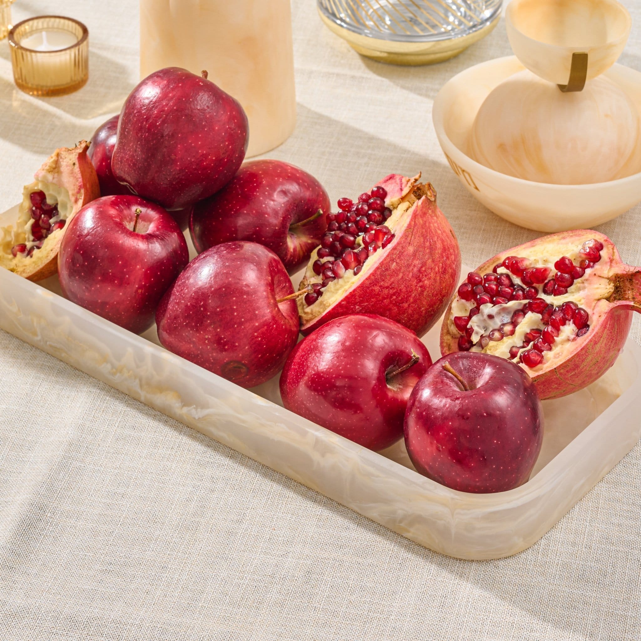 A rectangular resin tray with and cut open pomegranates, set on a light beige linen tablecloth with wooden bowls and candleholders in the background.