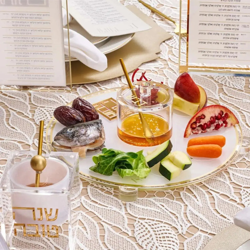 Rosh Hashanah Classic 2.0 tablescape with pomegranate, apple, and honey in glass dishes on lace tablecloth with prayer books and holiday placecards.