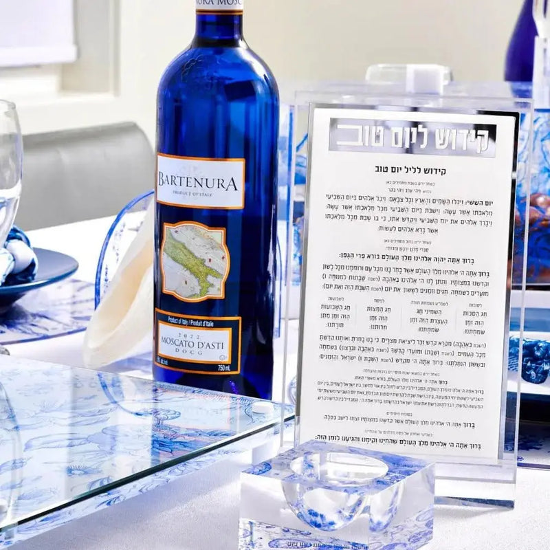 Shavuos French Toile 2024 tablescape featuring blue glass bottles, place cards, and elegant white dinnerware arranged on a table.