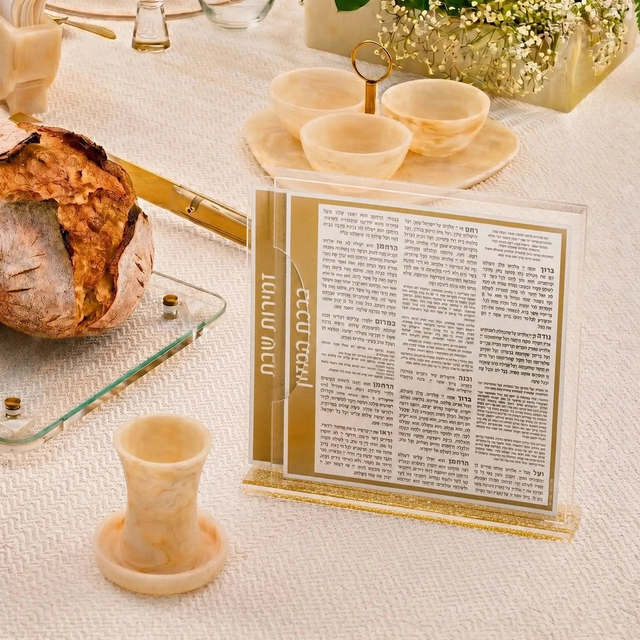 A clear acrylic tabletopBENCHERS styled on a white tablecloth with a loaf of bread, small marble cups, and a gold candlestick nearby.