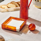 A set of amber marbled glass tableware on a white tablecloth, including a square napkin holder with white napkins, a resin amber pitcher  , and a small resin amber salt cellar