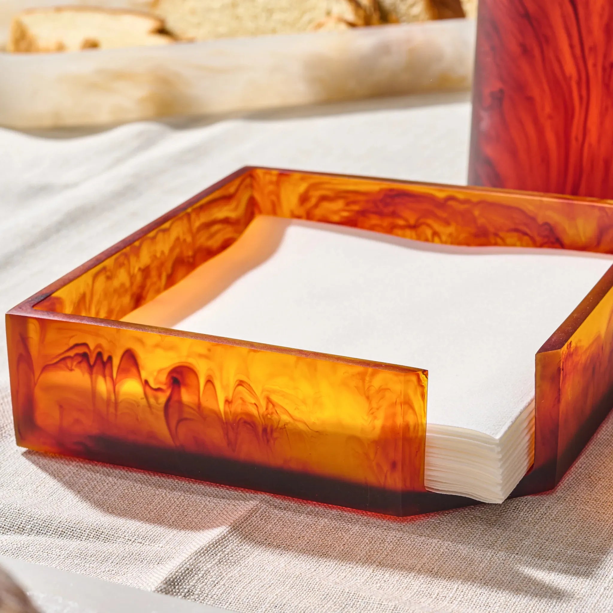 A square amber-orange resin napkin holder with a marbled swirl pattern, filled with white paper napkins on a light beige tablecloth.
