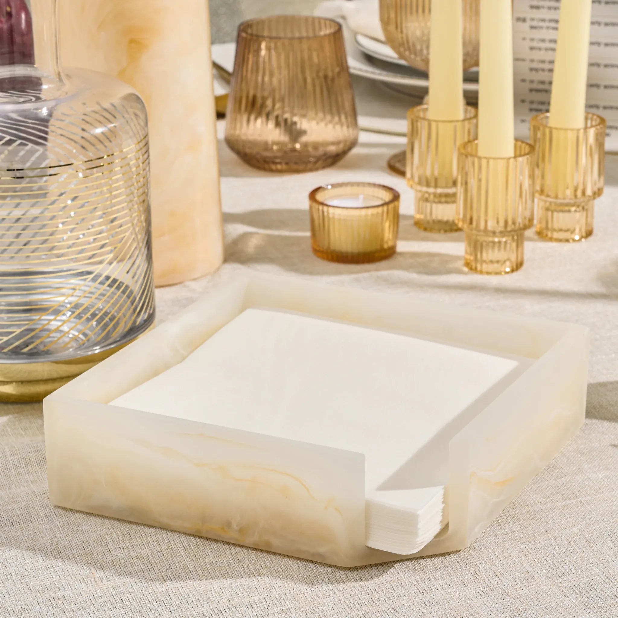 A square napkin holder made of cream-resinholding white folded napkins, styled on a linen tablecloth with candles and glassware in the background.