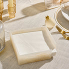A square napkin holder made of cream-resinholding white folded napkins, styled on a linen tablecloth with candles and glassware in the background.