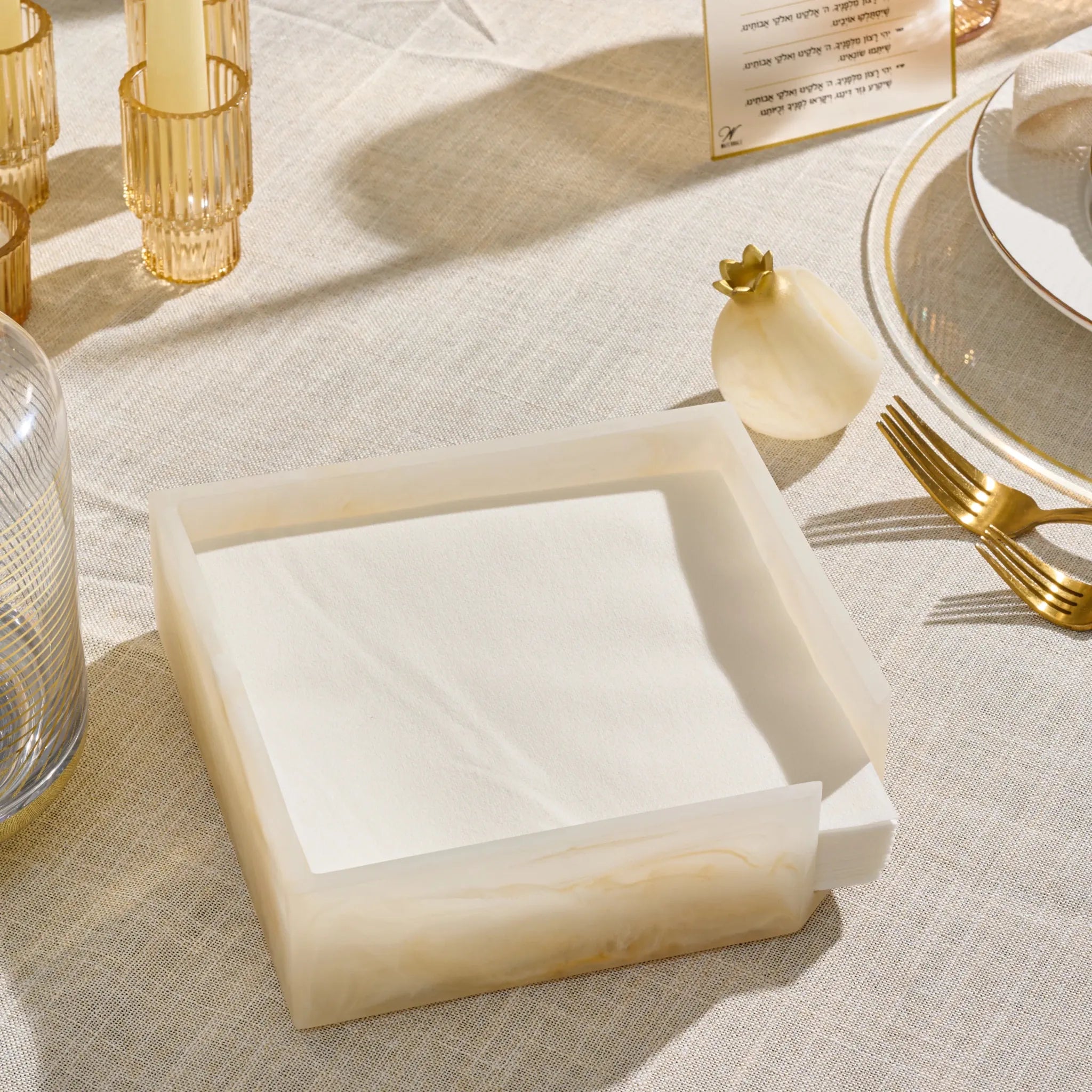 A square napkin holder made of cream-resinholding white folded napkins, styled on a linen tablecloth with candles and glassware in the background.