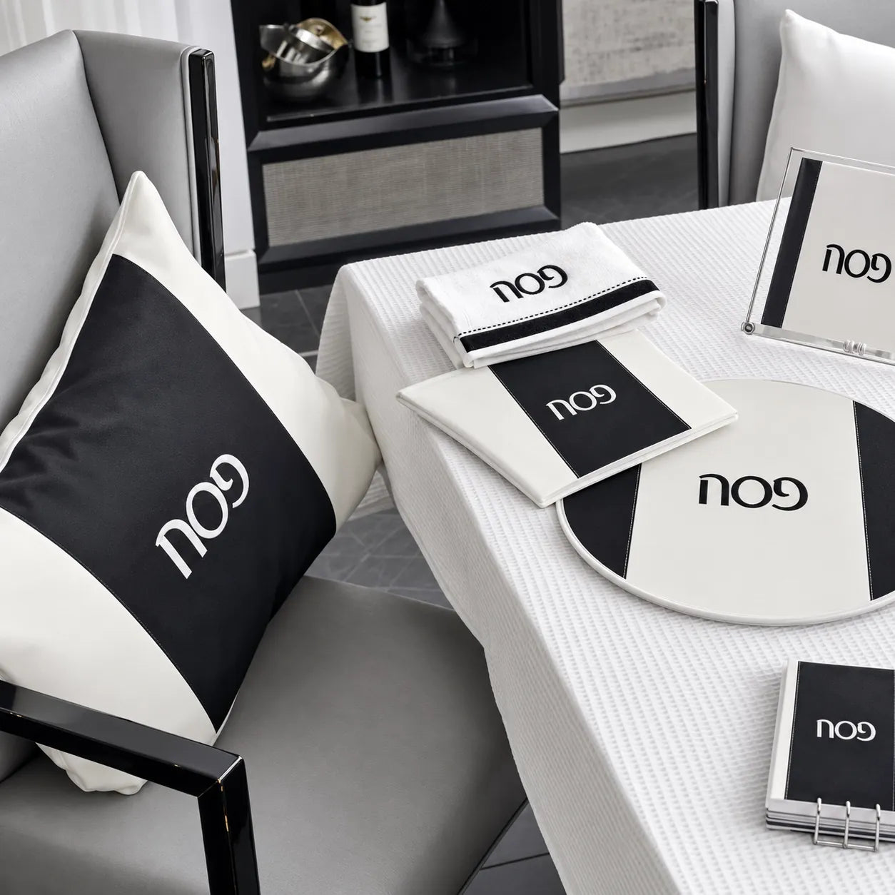 A styled interior scene showing coordinated black-and-whitePESACH SET  including a square throw pillow, folded napkins, placemat, sign, and notebook, arranged on a white tablecloth in a modern room.