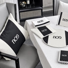A styled interior scene showing coordinated black-and-whitePESACH SET  including a square throw pillow, folded napkins, placemat, sign, and notebook, arranged on a white tablecloth in a modern room.