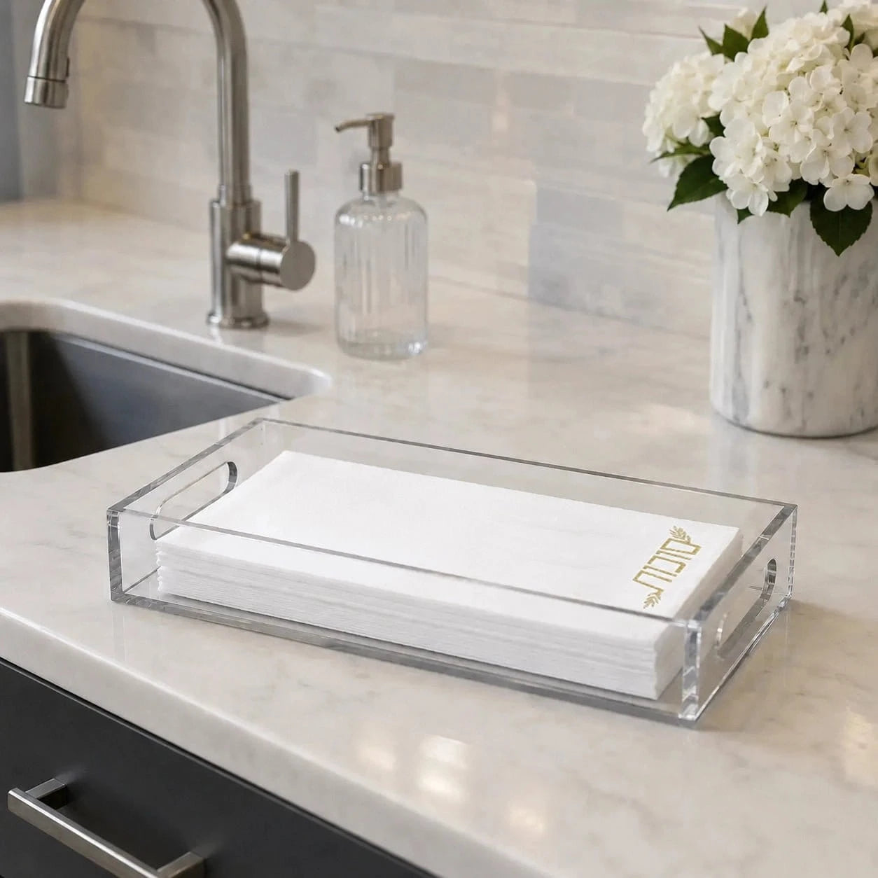 Clear acrylic rectangular tray holding a stack of white paper napkins with a small gold logo, placed on a light marble kitchen countertop near a sink and soap dispenser.