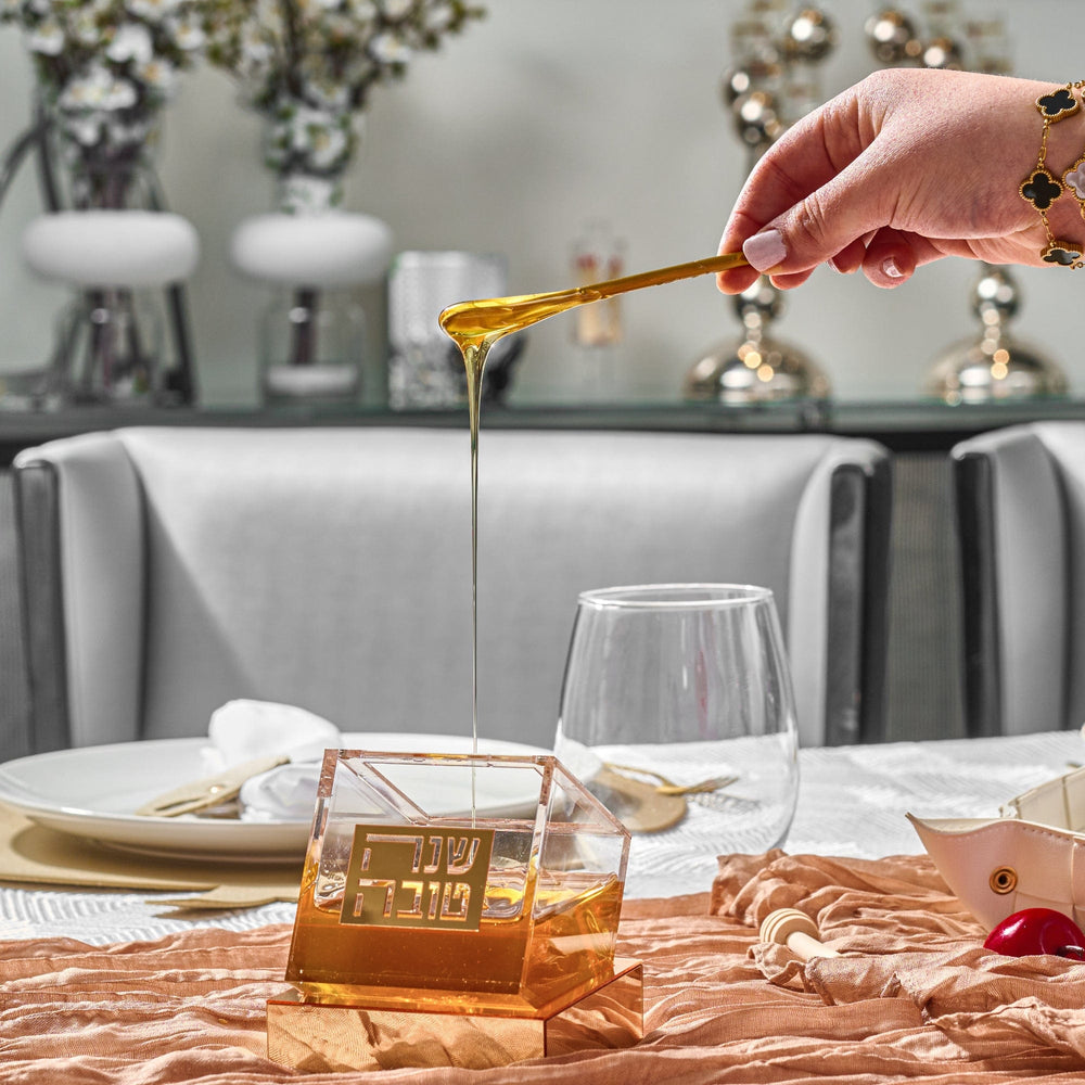 A stream of honey is pouring from a small, gold-colored spoon held by a woman's manicured hand into the Gold Tilt Honey Dish