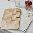 Woven challah board and cover set in natural tan with wooden square tiles, displayed with Shabbat candles and wine glasses on white surface.