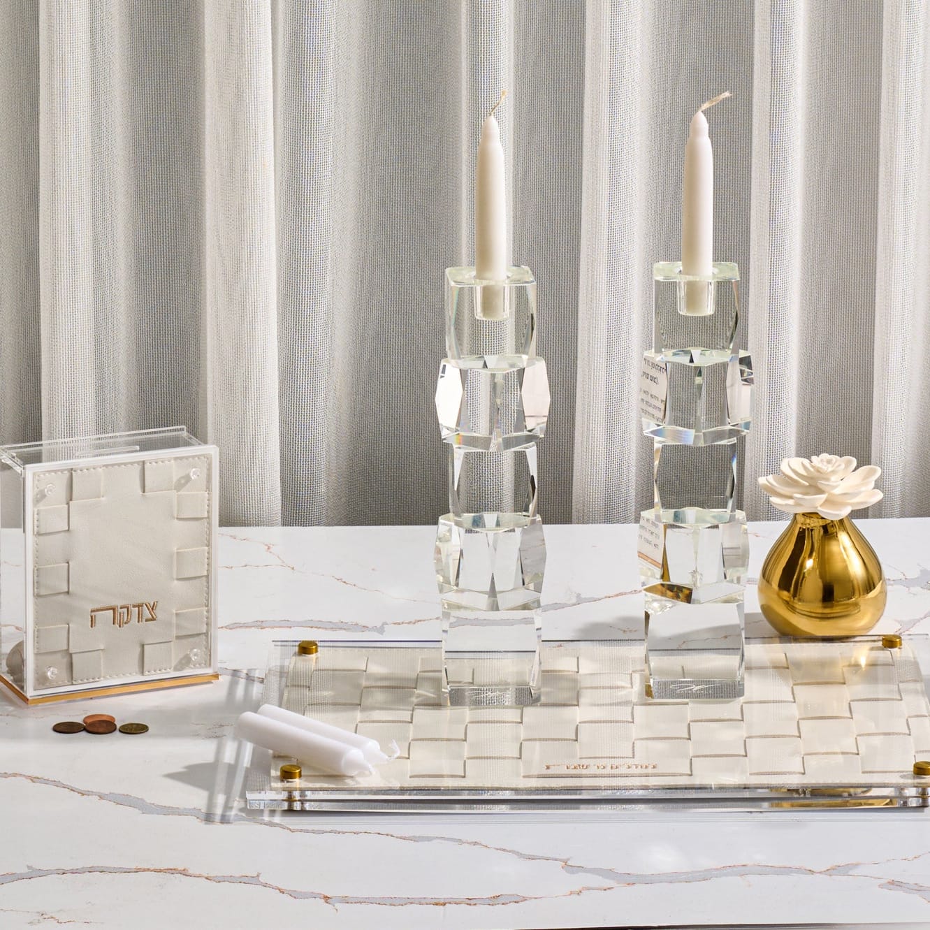 Woven hadlokas neiros set featuring tall crystal candle holders on a modern acrylic and woven leather tray with a matching tzedakah box and gold accents on a marble table.